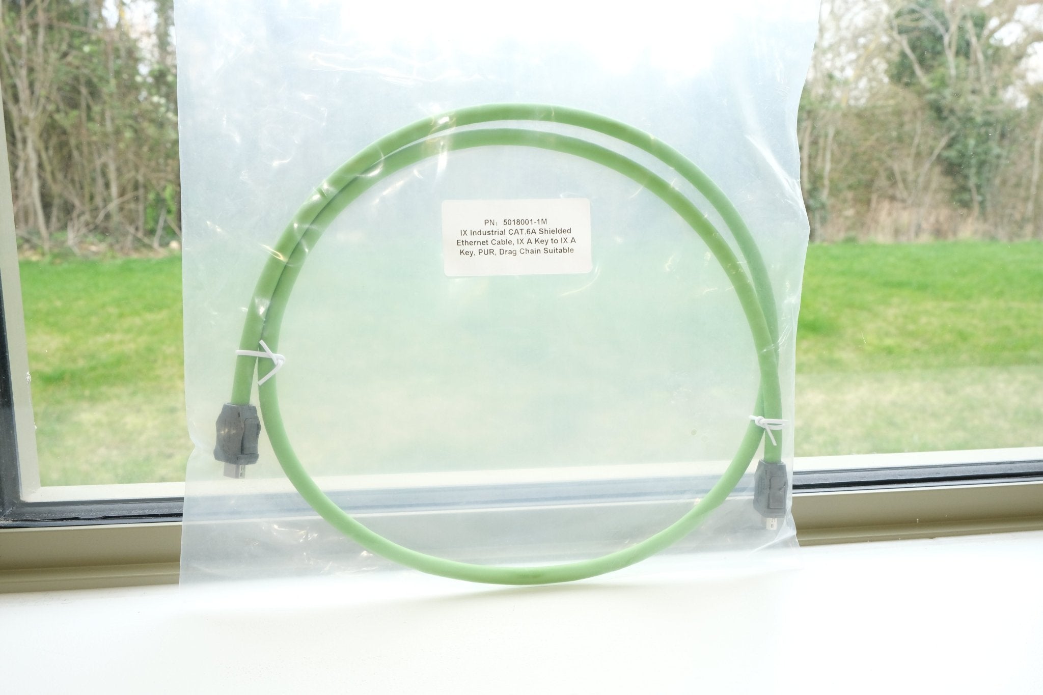 Green circular object in a clear plastic bag with a label, placed on a windowsill with a view of trees and grass outside.