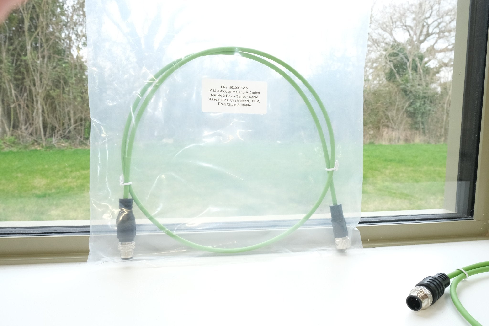 Green cable with connectors on a white surface near a window with a view of trees and grass.
