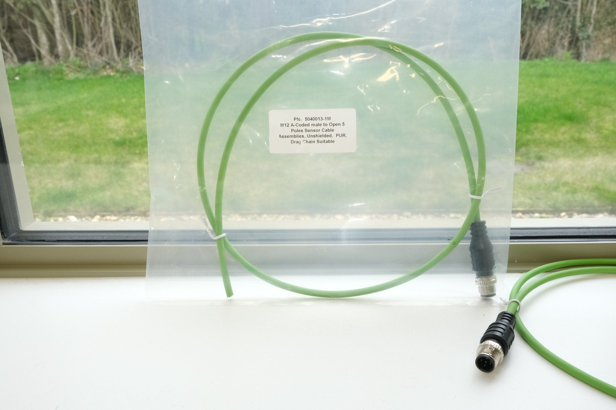 Green cable with connectors on a white surface, with a window in the background showing grass and trees.