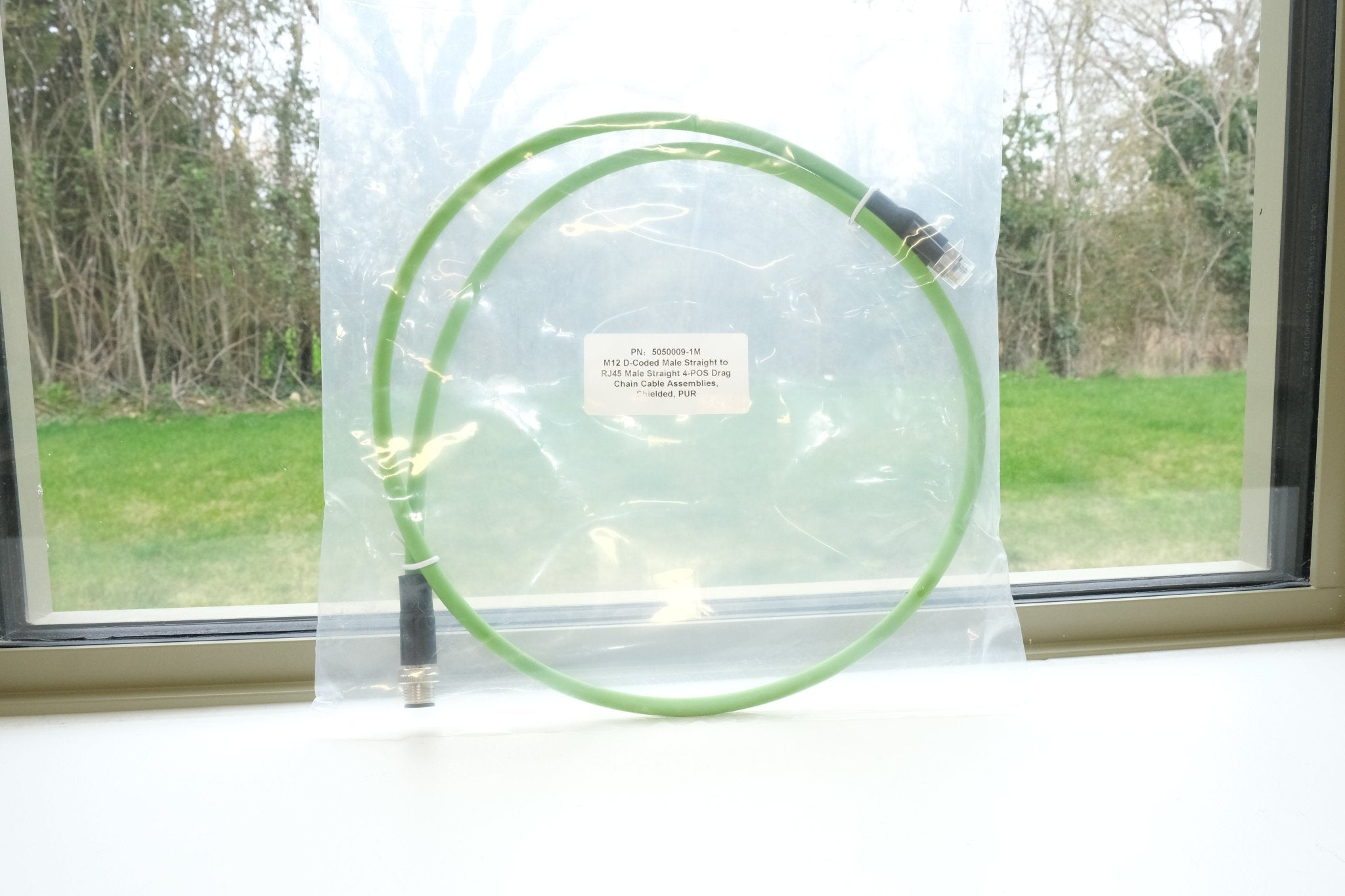 Green garden hose with nozzle on a window sill with a view of trees and grass outside.