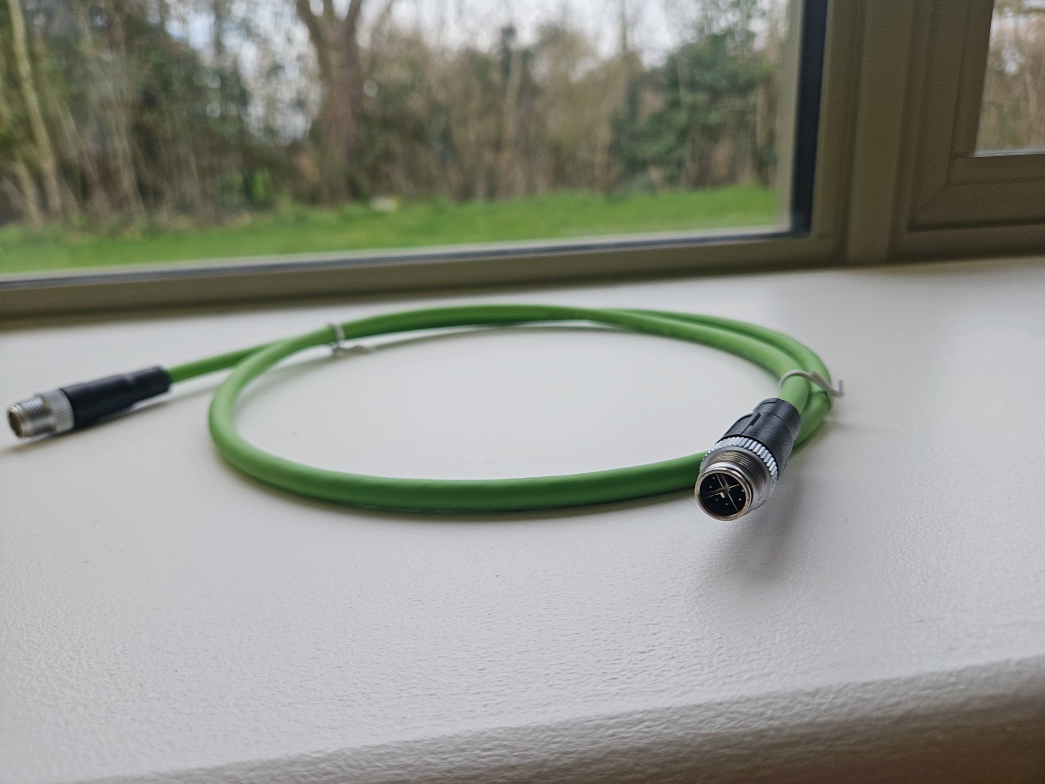 Green cable with connectors on a white surface near a window with a view of trees.