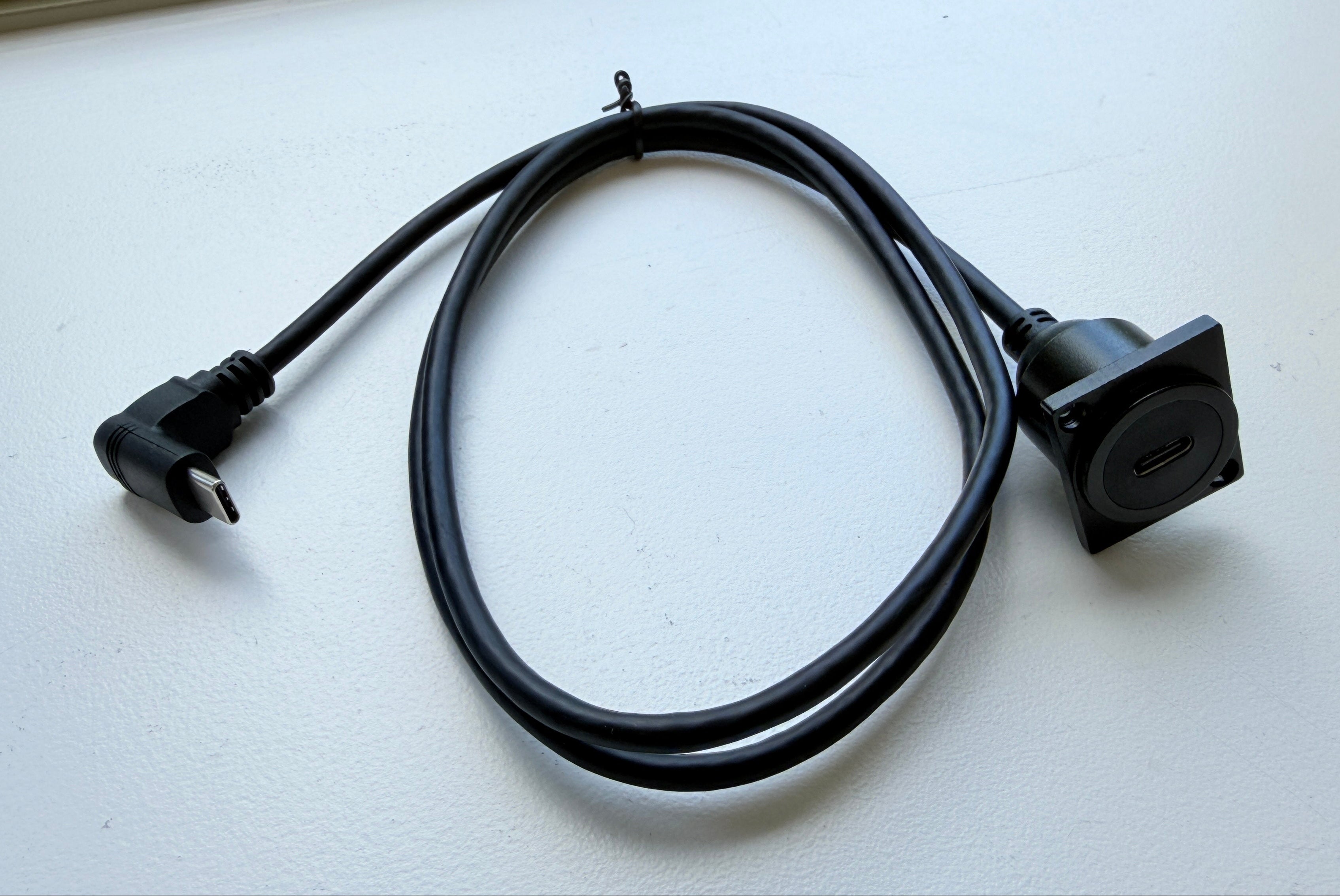 A black power adapter cable with a European plug on one end and a square connector on the other, coiled on a white surface.
